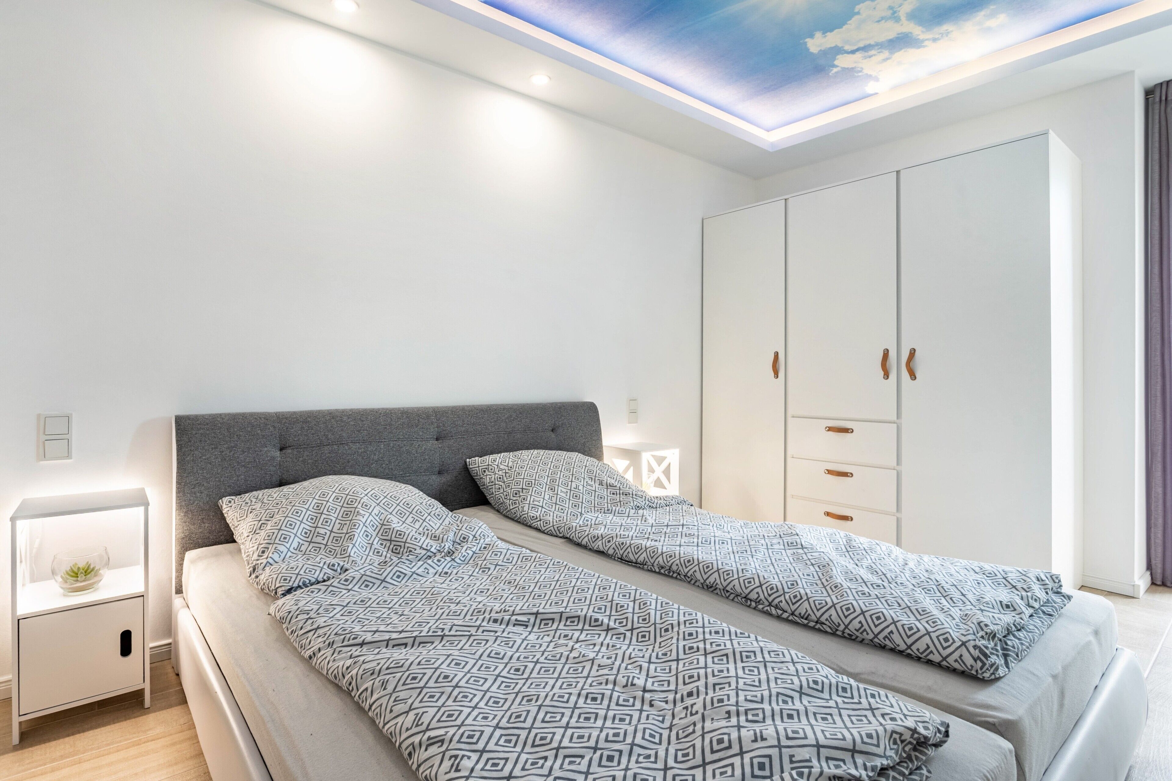 Ein modernes Schlafzimmer mit Bett, Nachttischen, einem Kleiderschrank und einer ansprechend gestalteten Deckenleuchte.