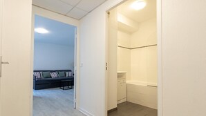 Ein Flur, der zu einem Badezimmer mit Dusche, Toilette und Waschbecken führt. Im angrenzenden Raum befindet sich eine Couch mit Kissen.