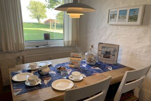 Ein Esstisch mit Geschirr, Tassen und einer Karaffe, ein Fenster mit Vorhängen und ein gerahmtes Bild an der Wand.
