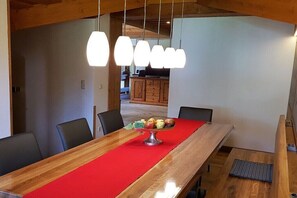 Ein Essbereich mit einem langen Holztisch, einer roten Tischläufer und einer Obstschale. Vier Stühle, ein Holzschrank im Hintergrund und hängende Pendelleuchten sind ebenfalls zu sehen.