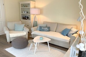 Ein Wohnzimmer mit einer Couch, zwei Sesseln, einem kleinen runden Tisch, einer Lampe und einem Bücherregal.