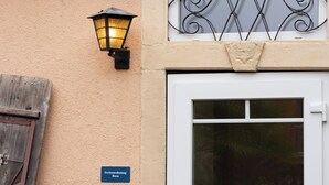 En bygnings facade med en beige væg, en trædør, en tændt væglampe, et vindue med dekorativt jernarbejde og et skilt med teksten 'Feriendwung Rosa'.