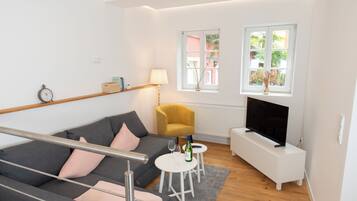 Ein modernes Wohnzimmer mit grauem Sofa, einem gelben Sessel, einem Fernsehgerät auf einem weißen Schrank und einem Fenster mit Blick auf Grünflächen.