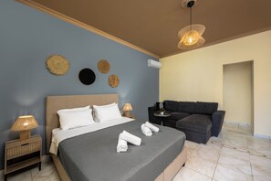 A neatly made bed with white linens, a dark gray headboard, and a bedside lamp. There is a dark gray sofa and a small round table with a lamp. - Pinotsi Room 6- ground floor (Simi)