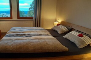 Ein Doppelbett mit gestreifter Bettwäsche und zwei Kissen, ein Nachttisch mit Lampe sowie ein Fenster mit Blick auf Bäume.