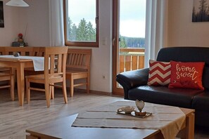 Ein gemütliches Wohnzimmer mit einem Holzesstisch, Stühlen, einer schwarzen Couch mit rotem Kissen und Blick auf Bäume durch das Fenster.
