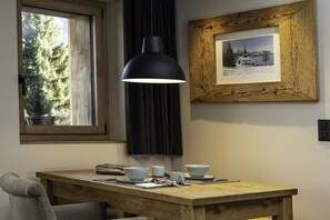 A dining area with a table, chairs, a pendant light, a framed picture, and a window with a view of trees. - Holiday apartment Sëléna (Bruson)