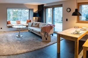 A modern living room with a sofa, a round table, a wooden dining table, and a large window with a view of trees. - Holiday apartment Sëléna (Bruson)