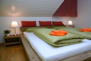 Ein Schlafzimmer mit schrägem Dach, zwei Nachttische mit Lampen, ein Bett mit grüner Bettwäsche und orangefarbenen gefalteten Gegenständen sowie roten Kissen.