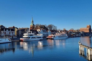 Ein Stadtbild am Wasser mit einem markanten Kirchturm, Booten, die an der Kaimauer festgemacht sind, und verschiedenen Gebäuden.