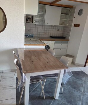A dining area with a wooden table and chairs, a kitchen with white cabinets and a tiled backsplash, and a clock on the wall. - Les Sables D'or N°28 - 2P6 (Agde)