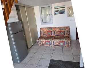 A kitchenette with a refrigerator, a patterned sofa, a window with a view, and a tiled floor. - Les Sables D'or N°28 - 2P6 (Agde)