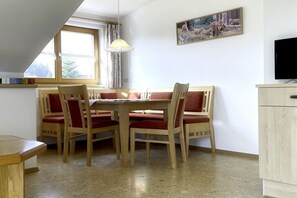 A dining area with a table and chairs, a window with curtains, a painting on the wall, and a television on a cabinet. - Holiday apartment with 2 bedrooms (Nesselwang)