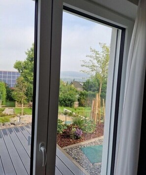 Blick durch eine Glastür auf einen Garten mit einer Holzterrasse, einem kleinen Teich und einem Gebäude in der Ferne.