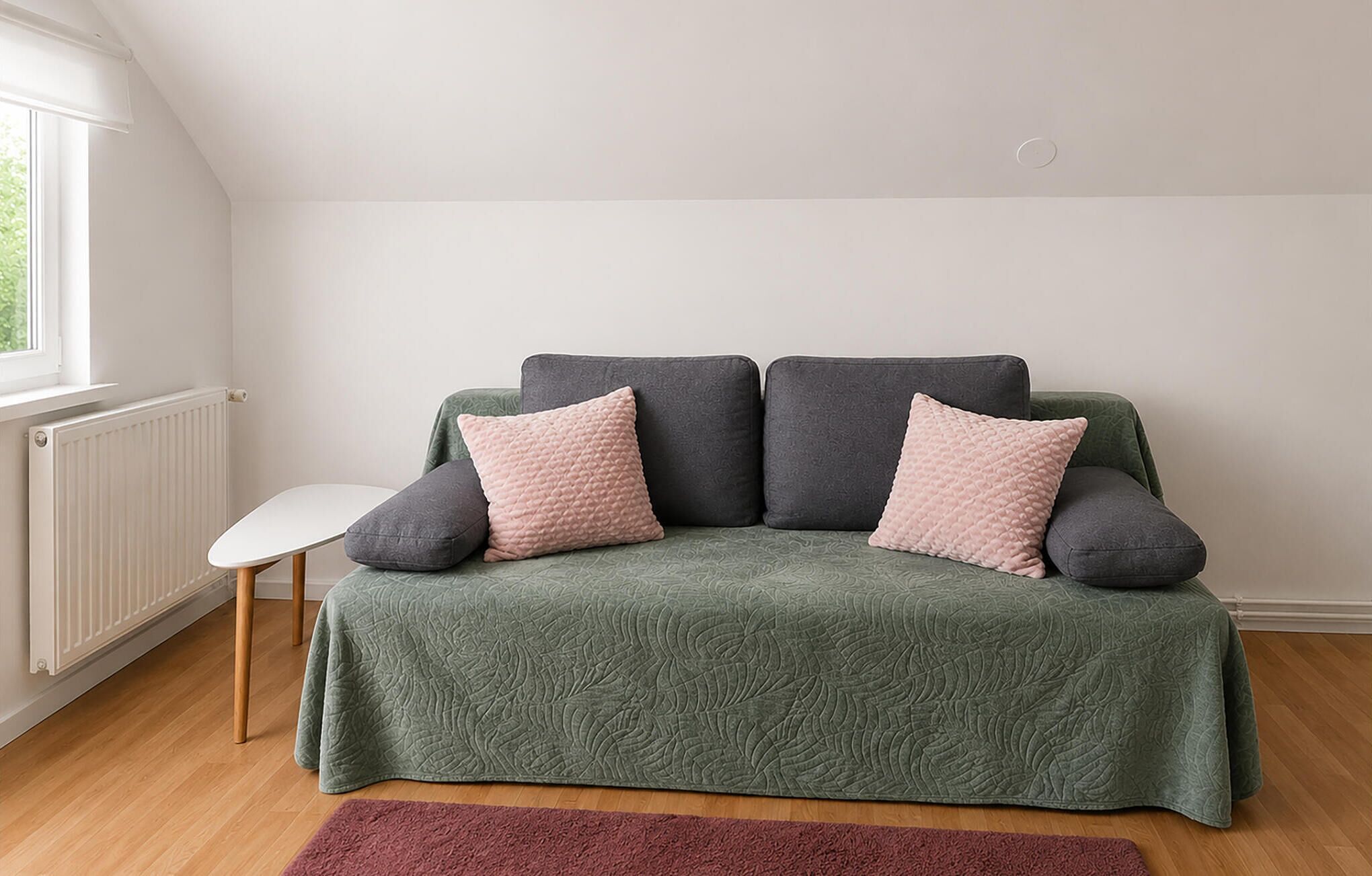 A room with a green sofa, two pillows, a white chair, a radiator, and a window.