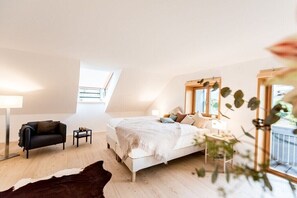 Ein Schlafzimmer mit schrägverdeckter Decke, einem Bett, einer kleinen Couch, einem Nachttisch und einem Fenster.