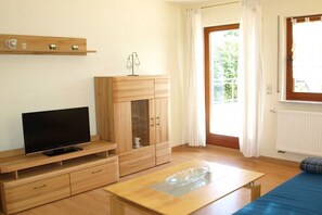 Ein Zimmer mit Fernseher auf einem Holzboden, einem Schrank mit Glastüren, einem Glastisch und einem blauen Sofa.