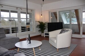 A modern living room with a sofa, a white armchair, a round coffee table, a television, and a large window. - Holiday apartment with 1 bedroom (Farchau)