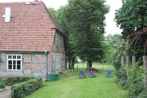 Ein traditionelles Backsteinhhaus mit einem Ziegeldach, einer Rasenfläche mit zwei Liegestühlen und einem großen Baum im Hintergrund.