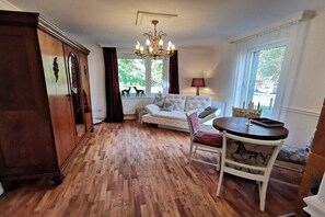 Ein Zimmer mit Holzboden, einem weißen Sofa, einem Esstisch mit Stühlen, einer Kristallleuchtre und einem fenster mit Vorhängen.