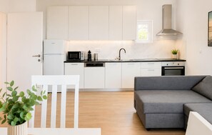 Eine moderne Küche mit weißen Schränken, einer Mikrowelle, einem Spülbecken und einem Herd. Im Wohnbereich befindet sich ein graues Sofa und ein weißer Sessel.