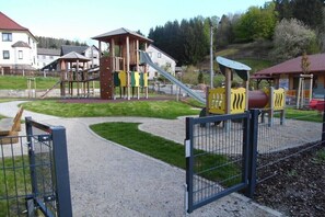 Ein Spielplatz mit Klettergerüsten und Rutschen, umgeben von einem Kiesweg und einem Metallzaun.