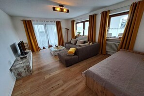 Ein Zimmer mit einer Couch, einem Bett, einem Fernseher und einem Vorhangfenster.