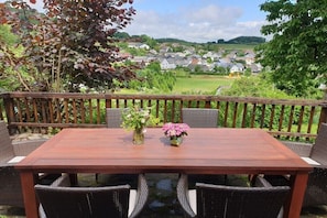 Eine Holzterrasse mit Tisch und Korbstühlen, mit Blick auf ein Dorf.