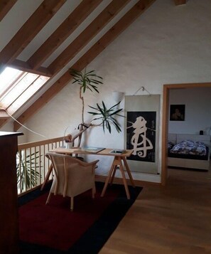 Ein Schlafzimmer im Loft-Stil mit schrägverdeckter Decke, einem Holztisch mit Stuhl, einer großen Pflanze und einem Bett mit gemustertem Überwurf.