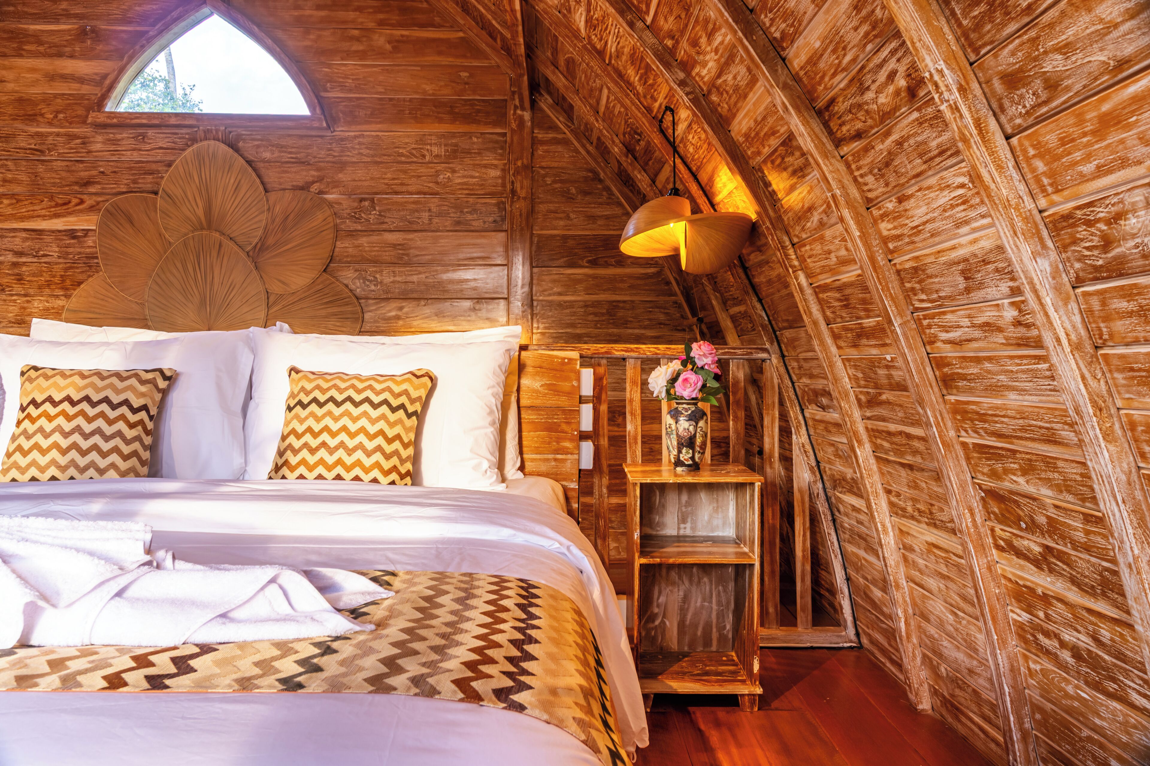 An attic bedroom with wooden walls, a bed with patterned bedding, a wooden nightstand, and a window.