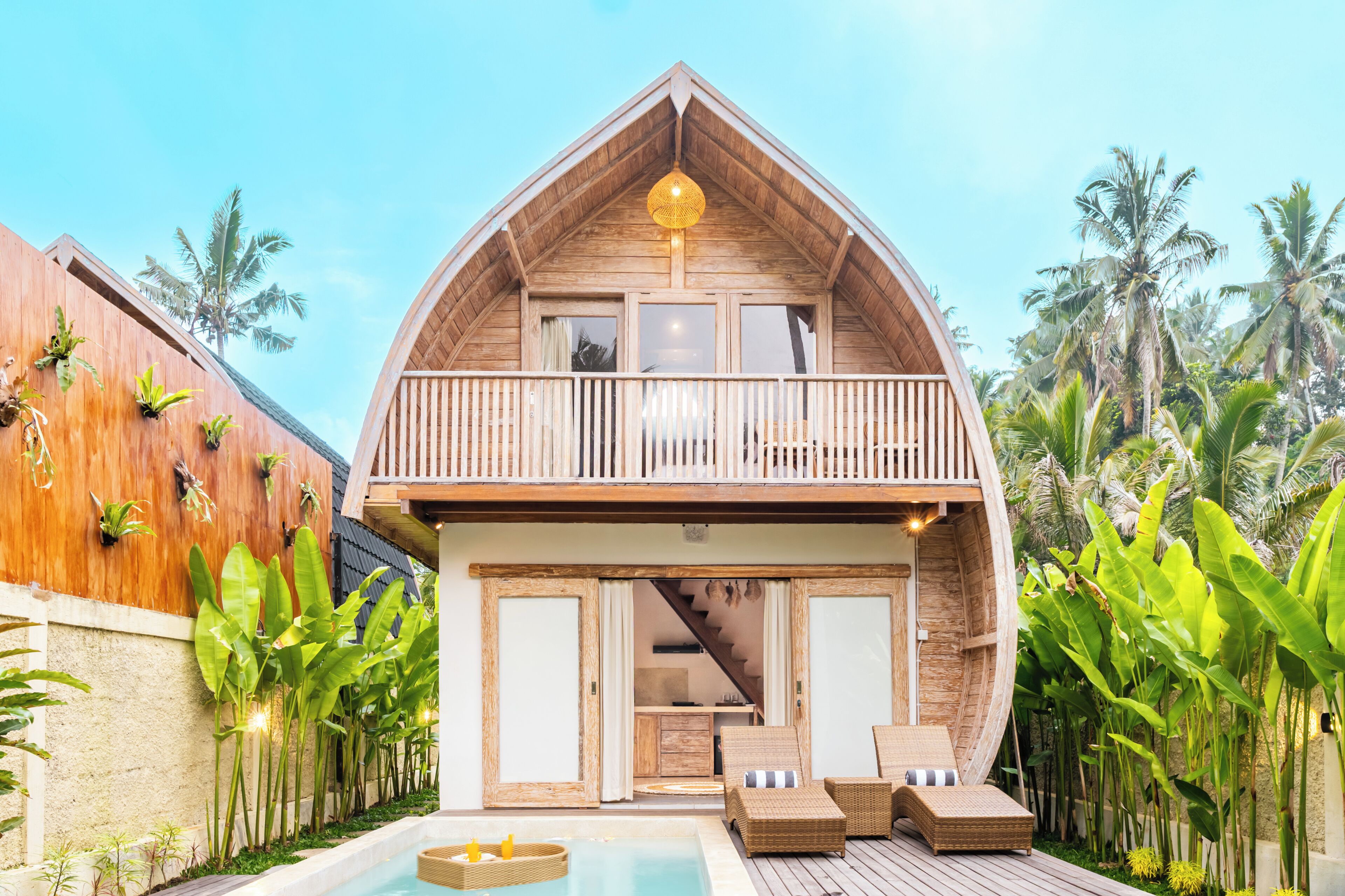 A two-story wooden house with a balcony, a pool, and tropical plants.