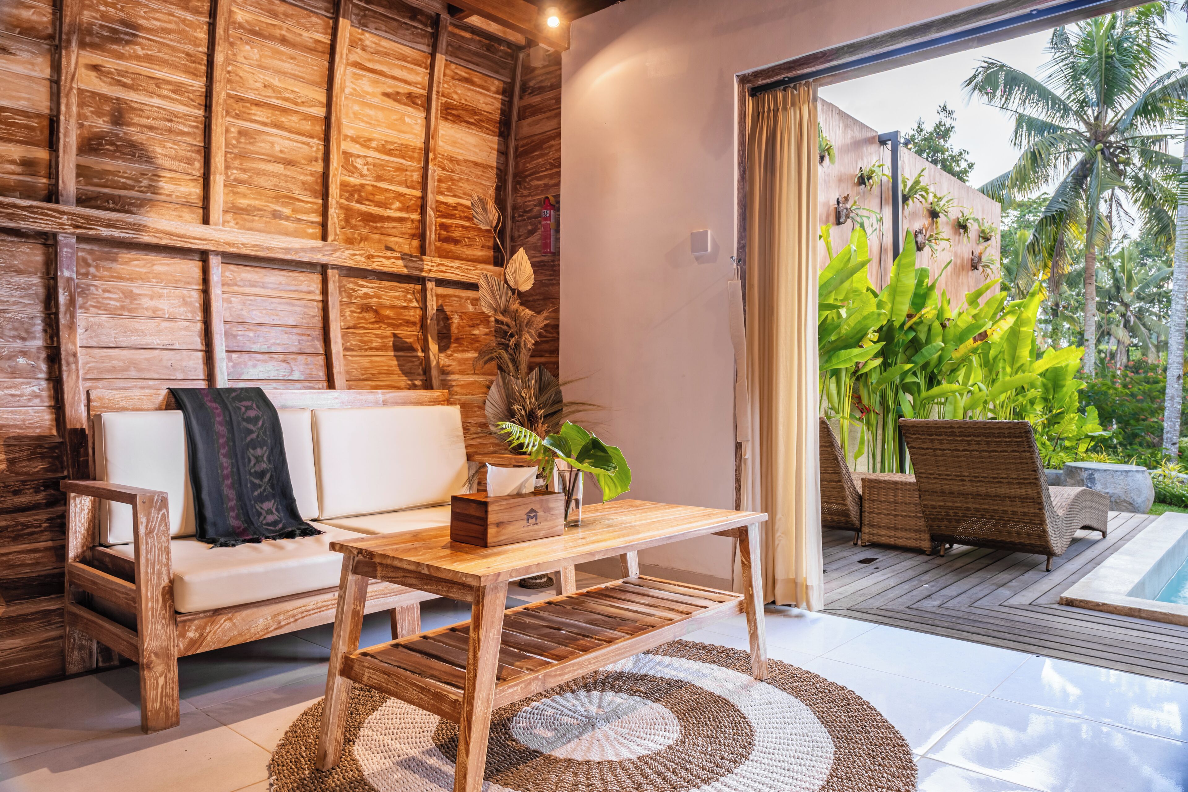 A modern living area with a wooden sofa, a wooden coffee table, and a view of a tropical garden through a glass door.