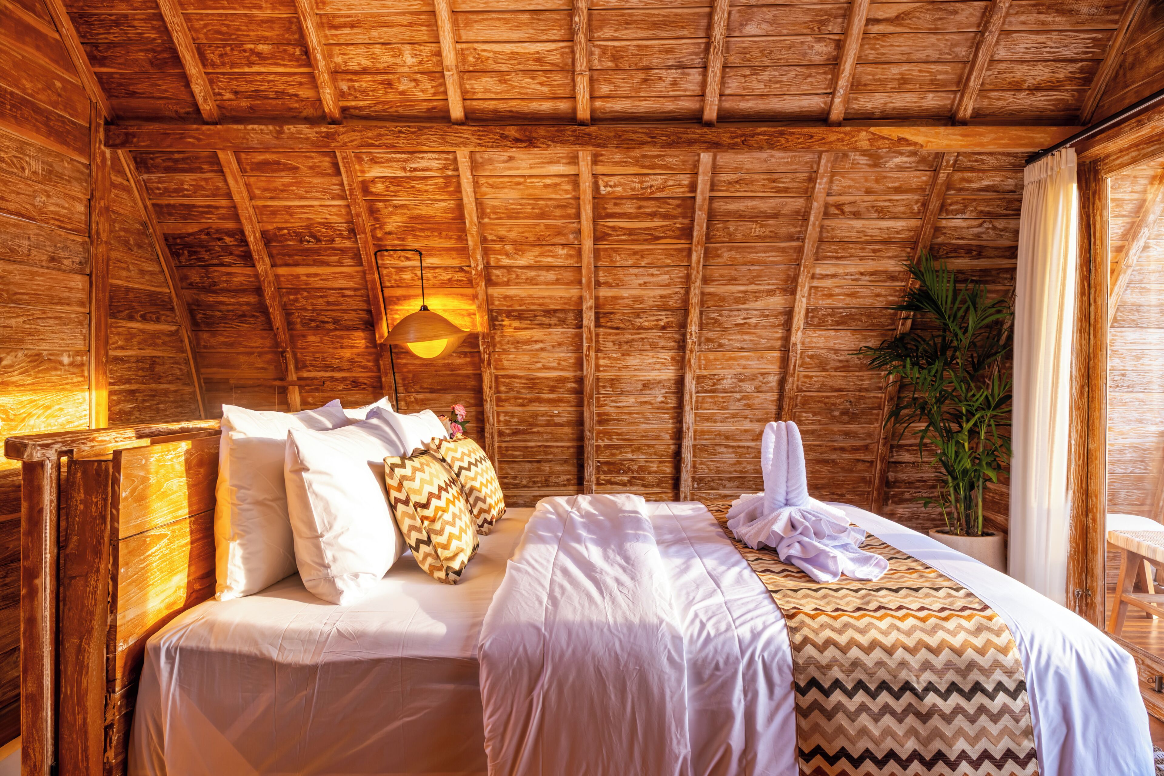 A wooden cabin room with two beds, a hanging light fixture, and a window with curtains.