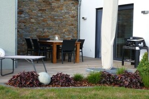 Eine Terrasse im Freien mit einer Steinmauer, einem Holztisch mit Stühlen, einem Grill und einer weißen Säule.
