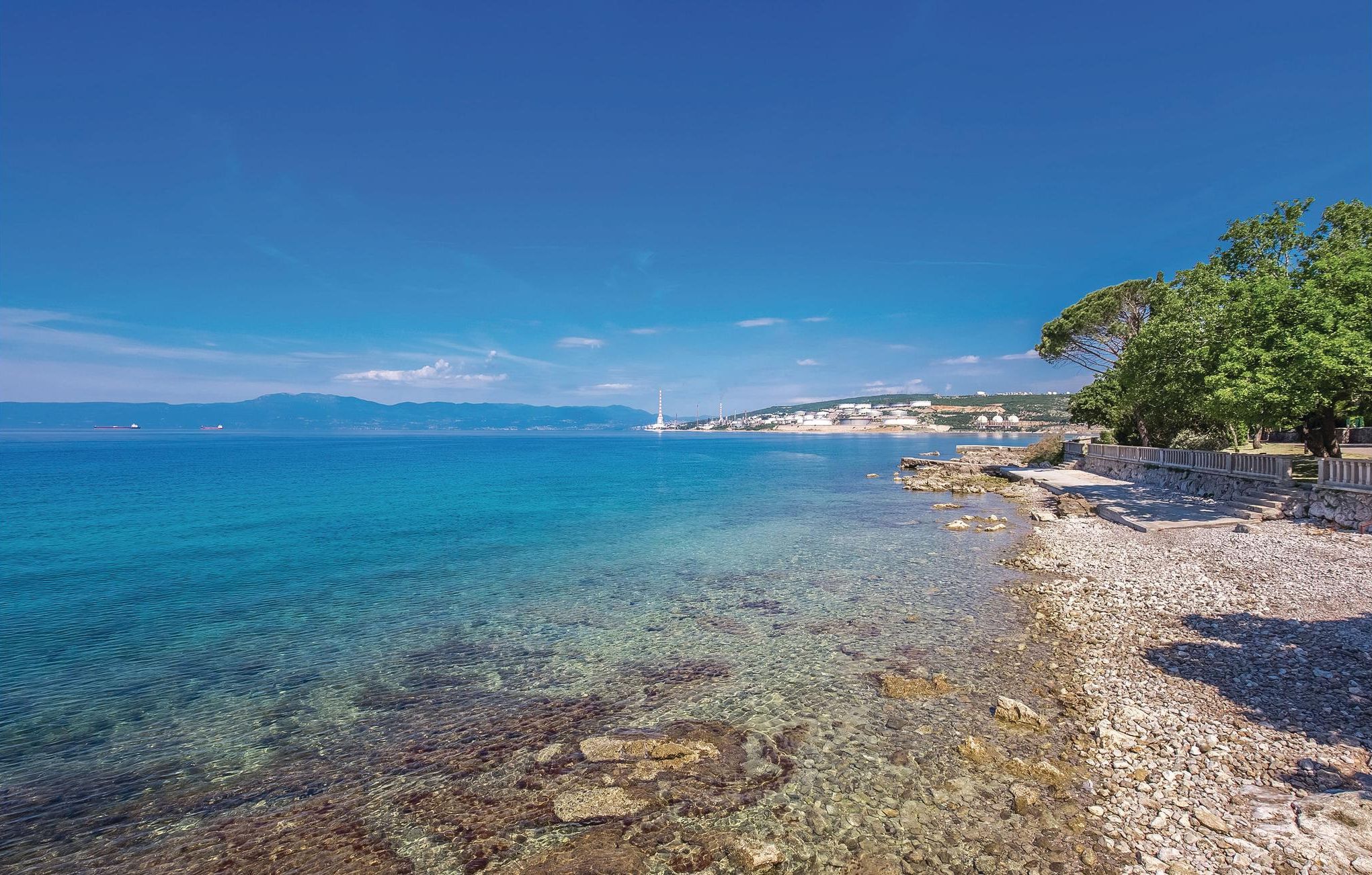 A coastal view with clear waters, a rocky shoreline, and distant buildings.
