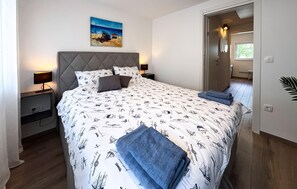 Ein Schlafzimmer mit einem Bett, Nachttischen, einem Kopfteil, einem Gemälde und einer Tür zu einem anderen Raum.