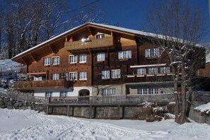 A multi-story wooden building with balconies, a stone wall, and a snowy landscape. - Holiday apartment with 2 bedrooms (Hasliberg Goldern)
