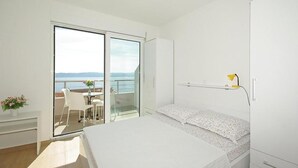A bedroom with a bed, a desk, a chair, a small table with flowers, a balcony with a view, and a nightstand. - Lovely apartment in Brela with WiFi (Brela)