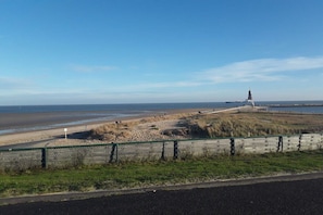 Paisagem costeira com um farol, dunas gramadas e uma estrada com cerca.