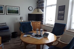 Ein Zimmer mit einem runden Holztisch, zwei blauen Sesseln, einem Sofa, einem Fernseher und gerahmten Bildern an den Wänden.