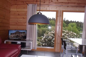 A wooden cabin interior with a TV, a red sofa, a dining table with chairs, and a large window with a view of trees and flowers. - Log cabin at the beginning (Thale-Friedrichsbrunn)