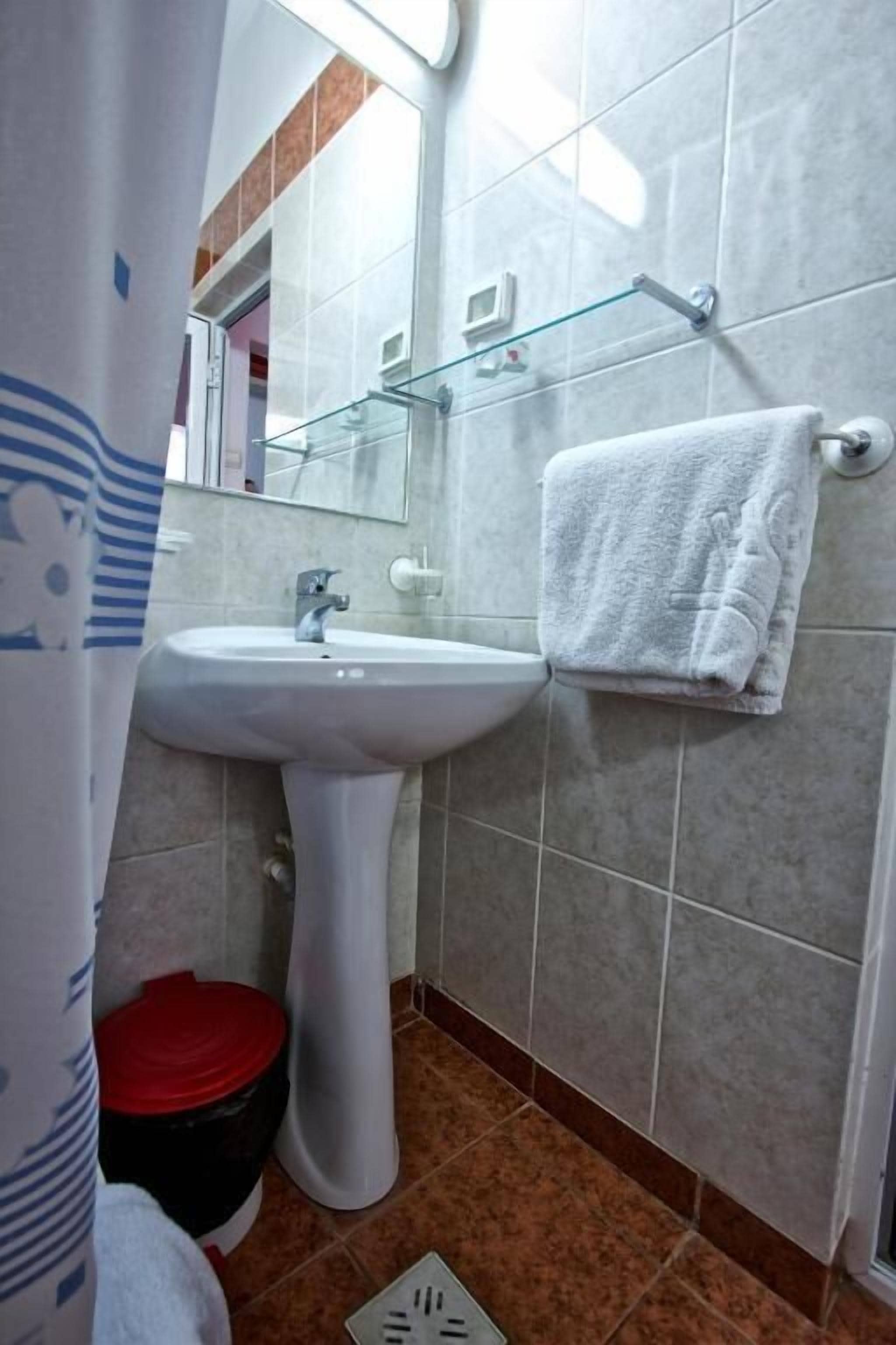 A bathroom with a white sink, a glass shelf, a towel hanging on the wall, and a red trash bin.