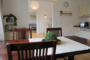 A dining area with a wooden table, chairs, a small potted plant, a white tablecloth, and a kitchen in the background. - Bender Grinding Shops (Ulsnis)
