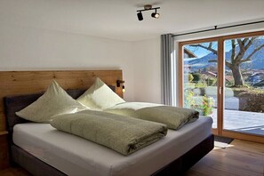 Ein Schlafzimmer mit Holzkopfstütze, einem großen Bett mit weißer Matratze, zwei Kissen und Blick auf eine gebirgige Landschaft durch eine Schiebetür.