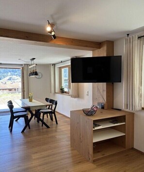 Ein modernes Wohnzimmer mit an der Wand montiertem Fernseher, einem Essbereich mit Tisch und Stühlen und Holzboden.