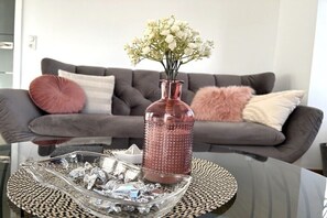 Ein Wohnzimmer mit einem grauen Sofa, einer rosafarbenen Vase mit weißen Blumen und einer Glasablage mit silbernen Gegenständen.