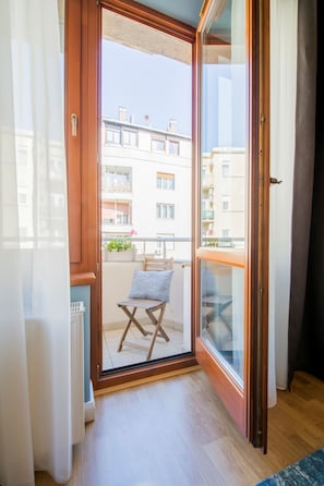 Ein Balkon mit einem Holzstuhl und einem kleinen Tisch, Blick auf benachbarte Gebäude und eine Tür zum Innenbereich.