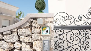 Un muro de piedra con un cartel de número de casa, un buzón de seguridad y una puerta metálica decorativa.