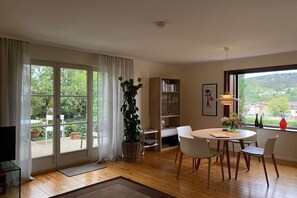 A room with a large sliding glass door, a dining table with chairs, a bookshelf, a potted plant, and a window with a view of greenery and buildings. - Holiday home with 1 bedroom (Bad Dürkheim)