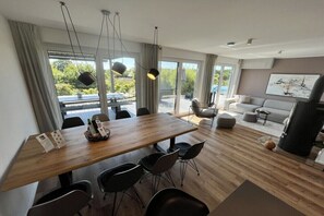 Ein modernes Wohnzimmer mit einem Holzesstisch, schwarzen Stühlen, einem grauen Sofa und einem großen Fenster mit Blick auf Grünflächen.